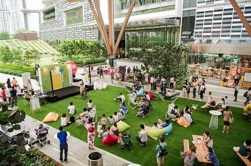 An outdoor event with a stage and audience seated on grass, beanbags, and in hammocks. Modern architecture visible in the background.